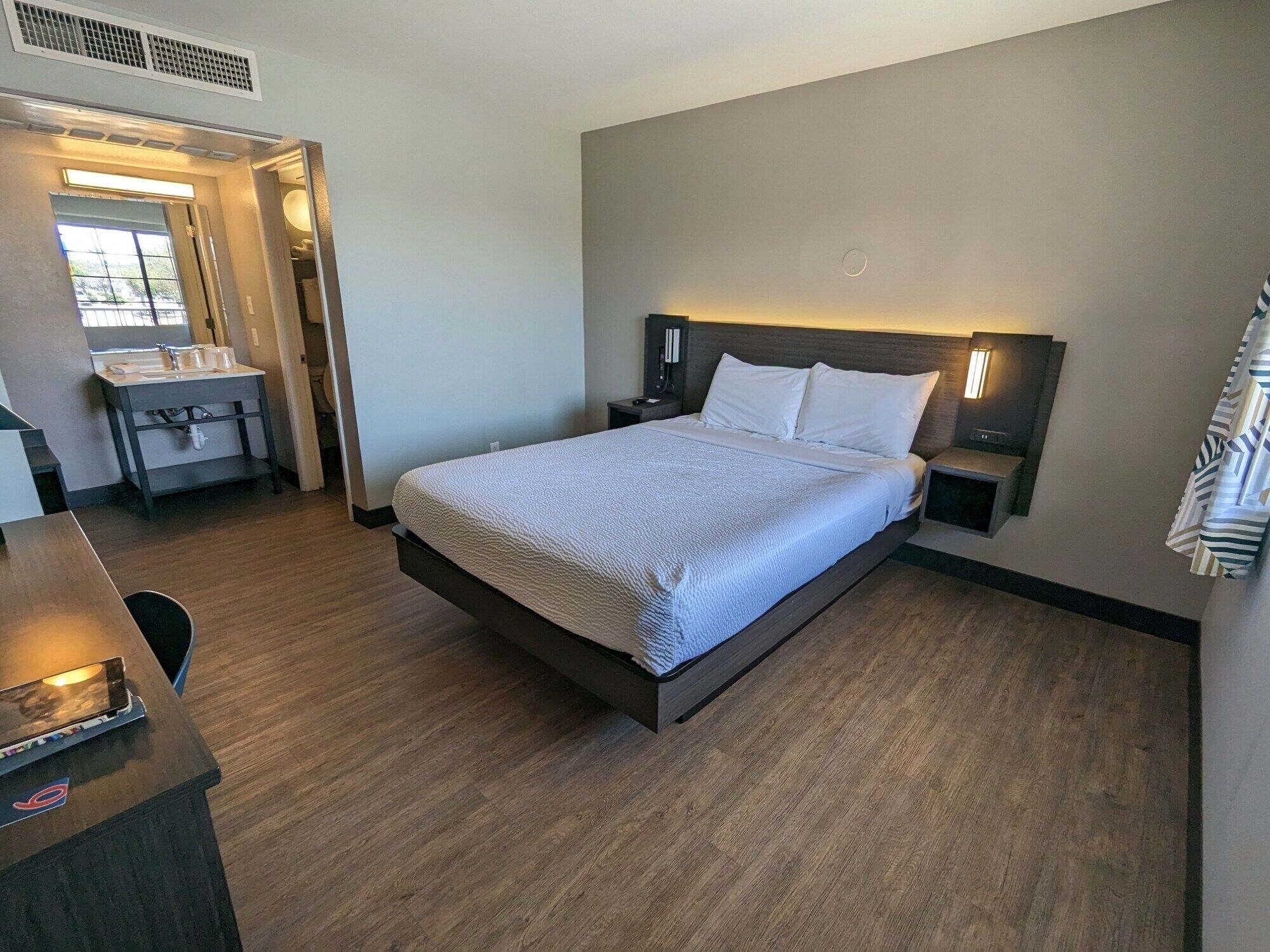A hotel room with a large bed, bedside tables, a desk with a chair, a window, and a bathroom with a sink and mirror.