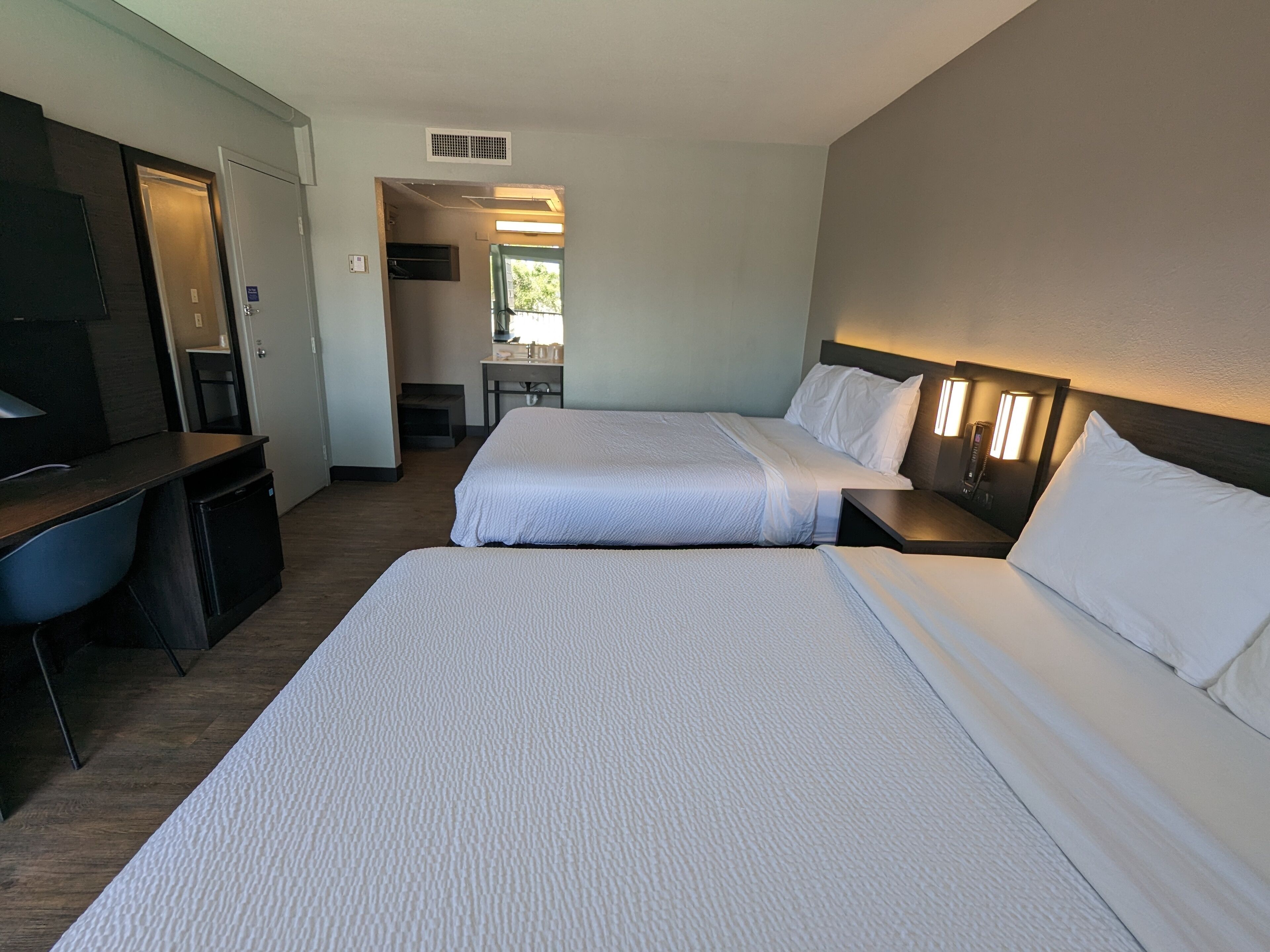 A hotel room with two beds, a desk, a chair, and a kitchenette.