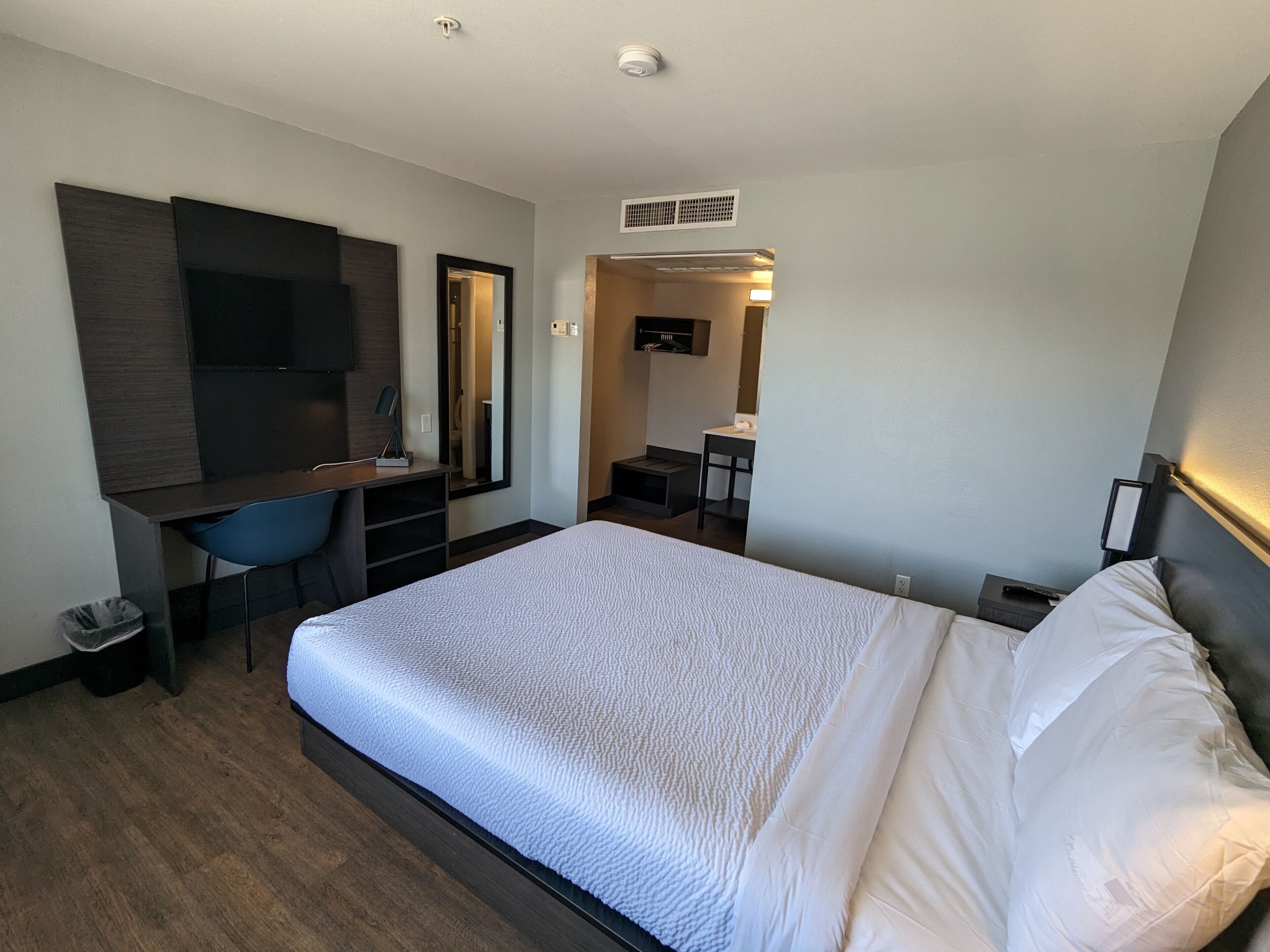 A hotel room with a large bed, a desk with a chair, a mirror, and a small kitchenette area.