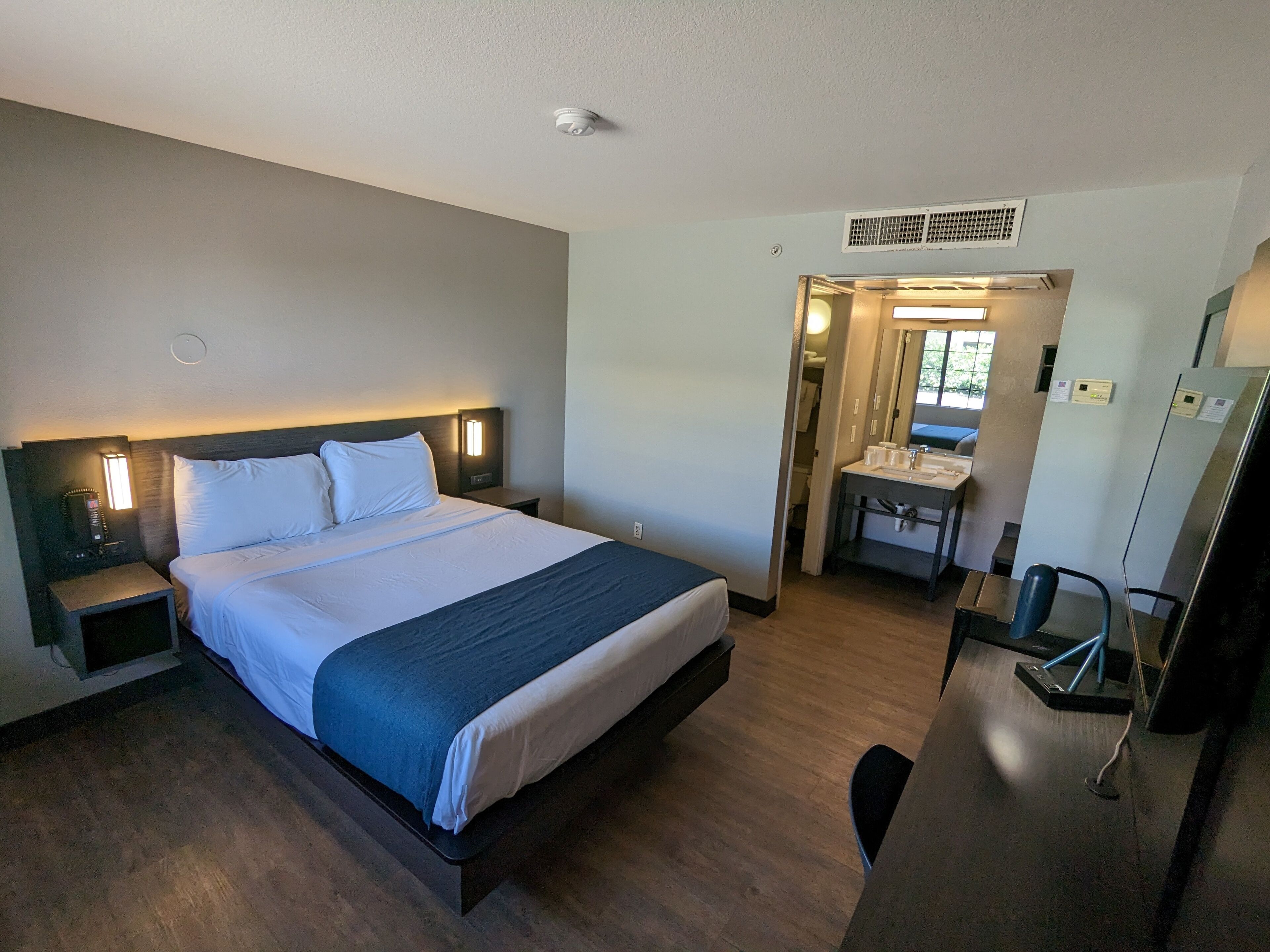 A hotel room with a bed, bedside table, lamp, and a bathroom with a sink and mirror.