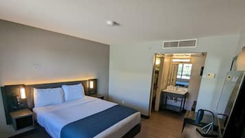 A hotel room with a large bed, a bedside table with a lamp, a desk with a chair, a mirror, and a small bathroom with a sink and toilet.