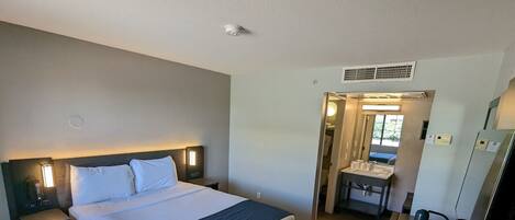 A hotel room with a large bed, a bedside table with a lamp, a desk with a chair, a mirror, and a small bathroom with a sink and toilet.