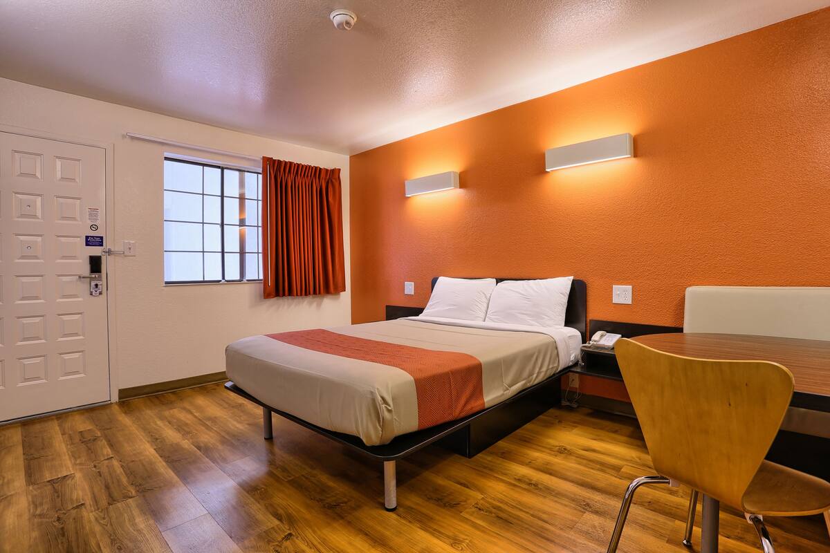 A hotel room with a bed, a desk, a chair, a window with curtains, and a wall-mounted light fixture.