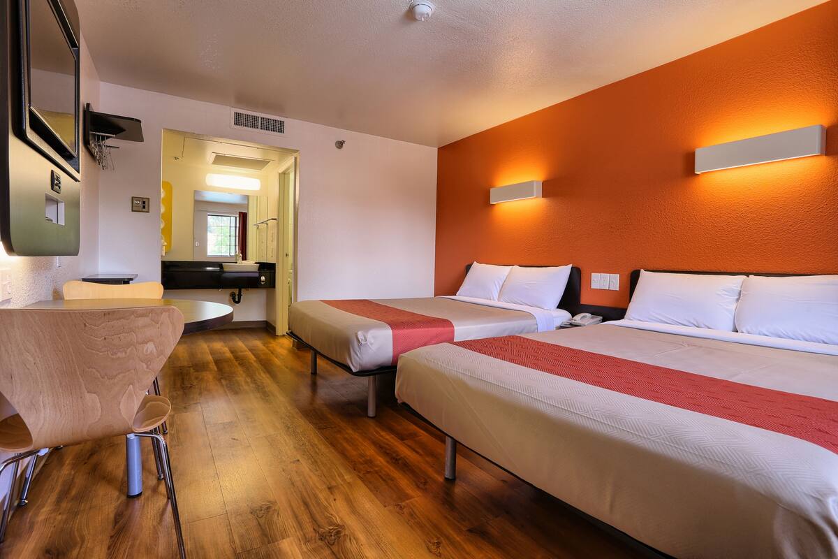 A hotel room with two beds, a wooden floor, a desk with a chair, and a bathroom visible through an open door.