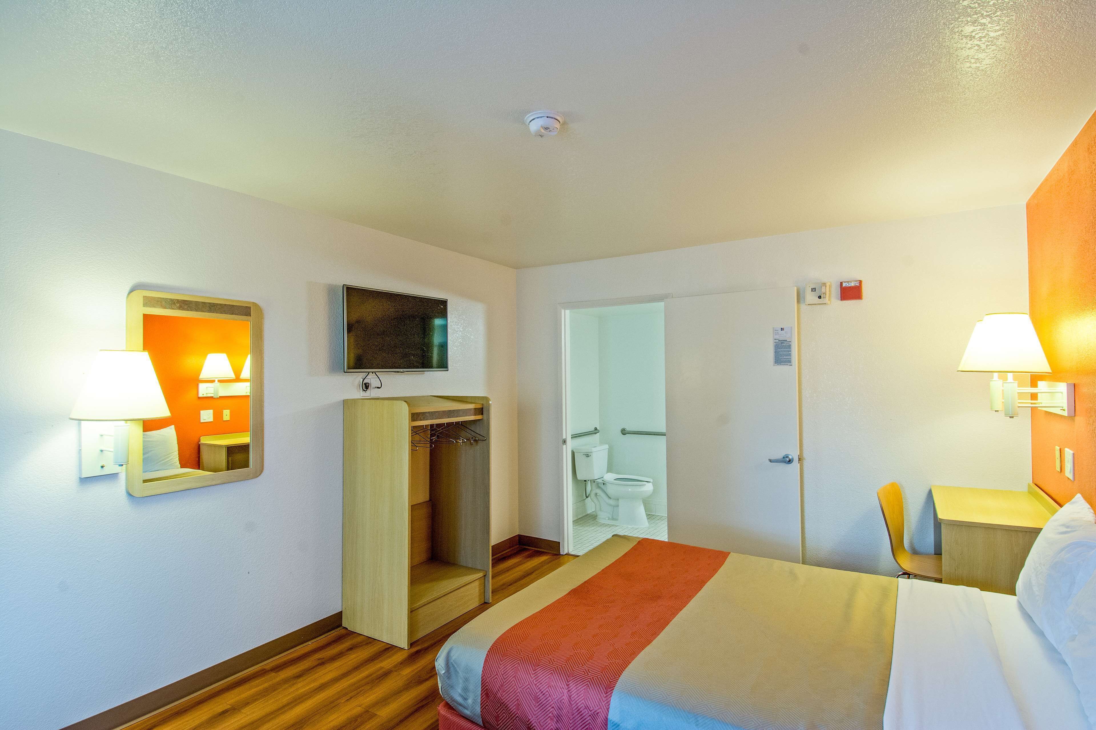 A hotel room with a bed, a wardrobe, a desk, a chair, a television, and a bathroom visible through an open door.
