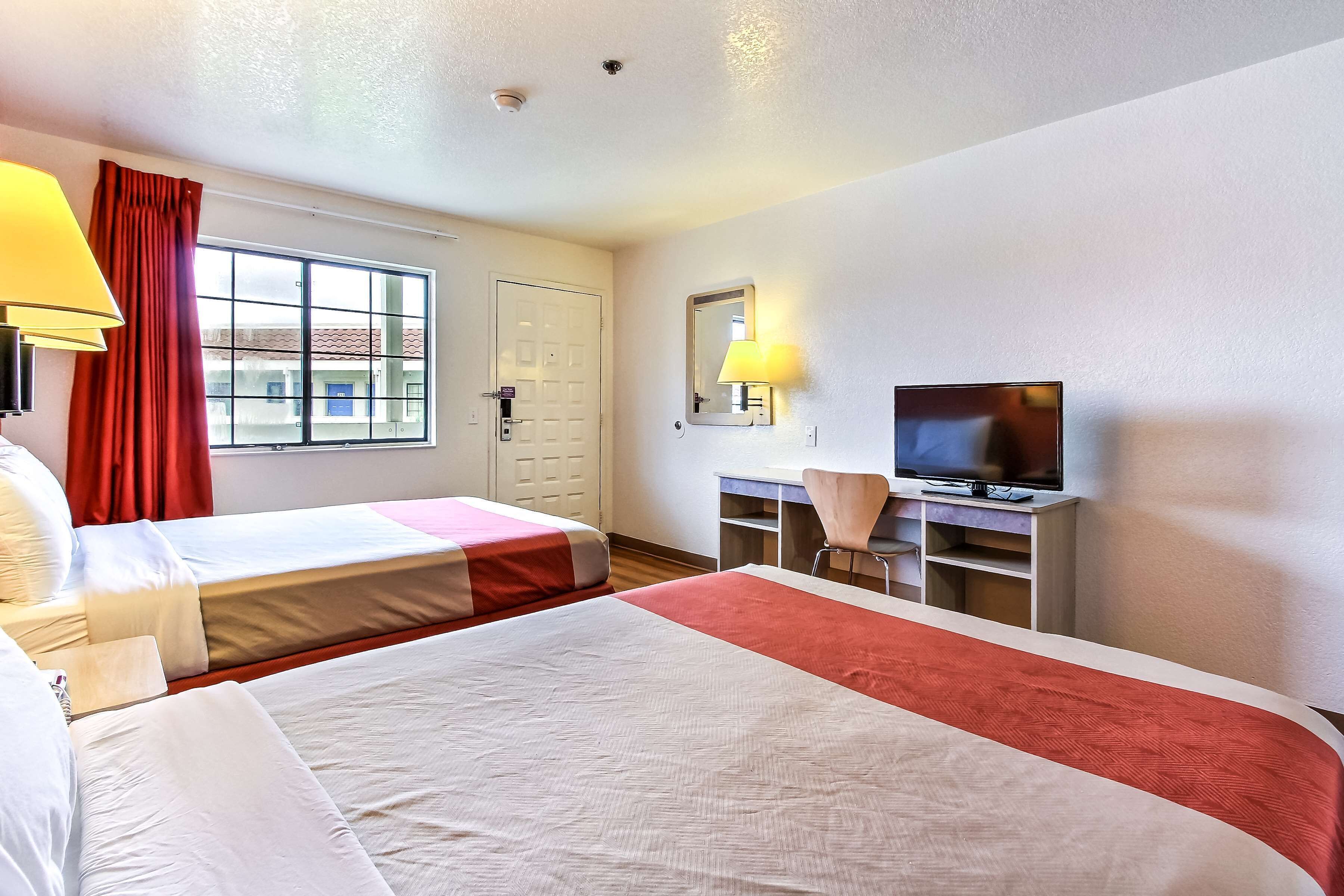 Une chambre d’hôtel avec deux lits, un bureau avec une télévision, une chaise et une fenêtre avec des rideaux rouges.
