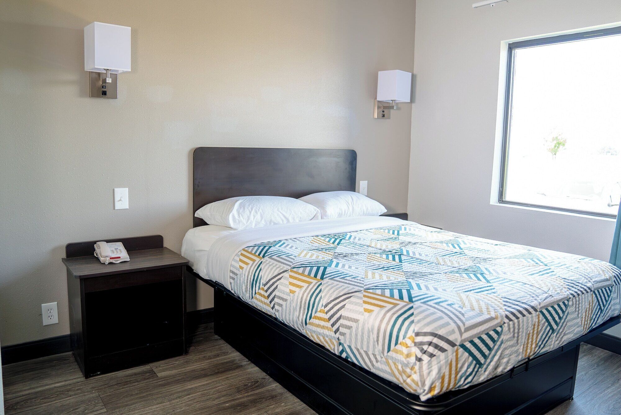 A hotel room with a bed, a bedside table with a phone, two wall-mounted lamps, and a window with a view.