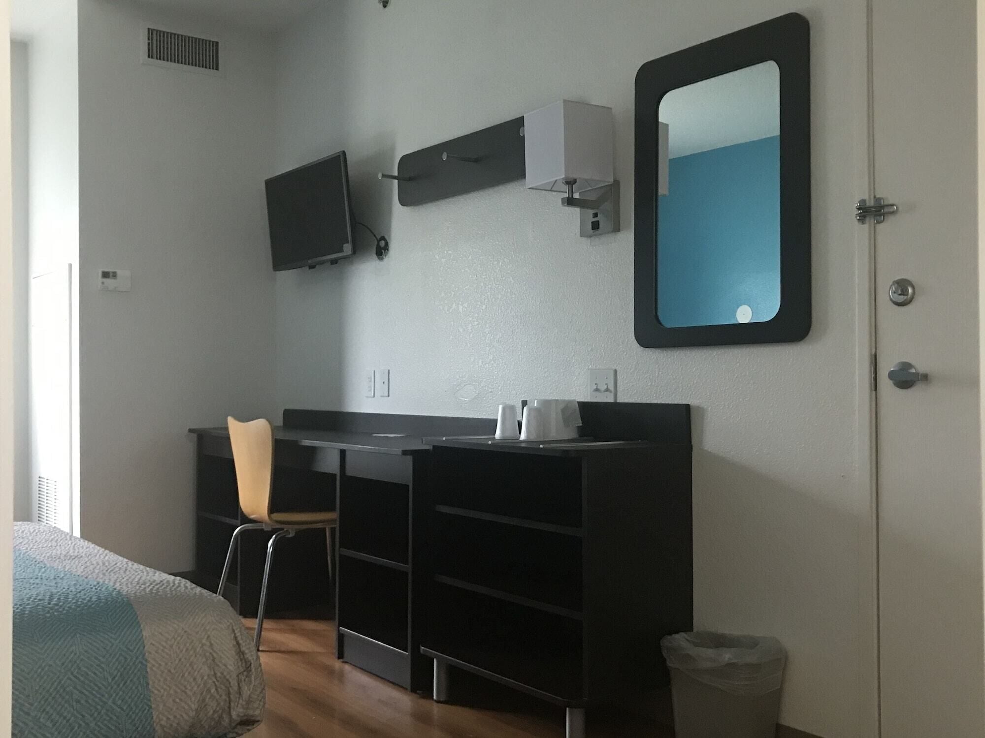 Una habitación pequeña y moderna con cama, escritorio, silla, televisor, espejo y cesto de basura.