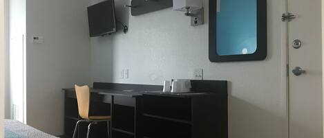 Una habitación pequeña y moderna con cama, escritorio, silla, televisor, espejo y cesto de basura.