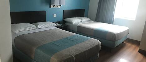 A hotel room with two beds, a nightstand, a wall-mounted lamp, and a window with curtains.