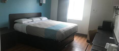 A hotel room with a bed, a desk with a chair, a window with curtains, and wall-mounted lamps.