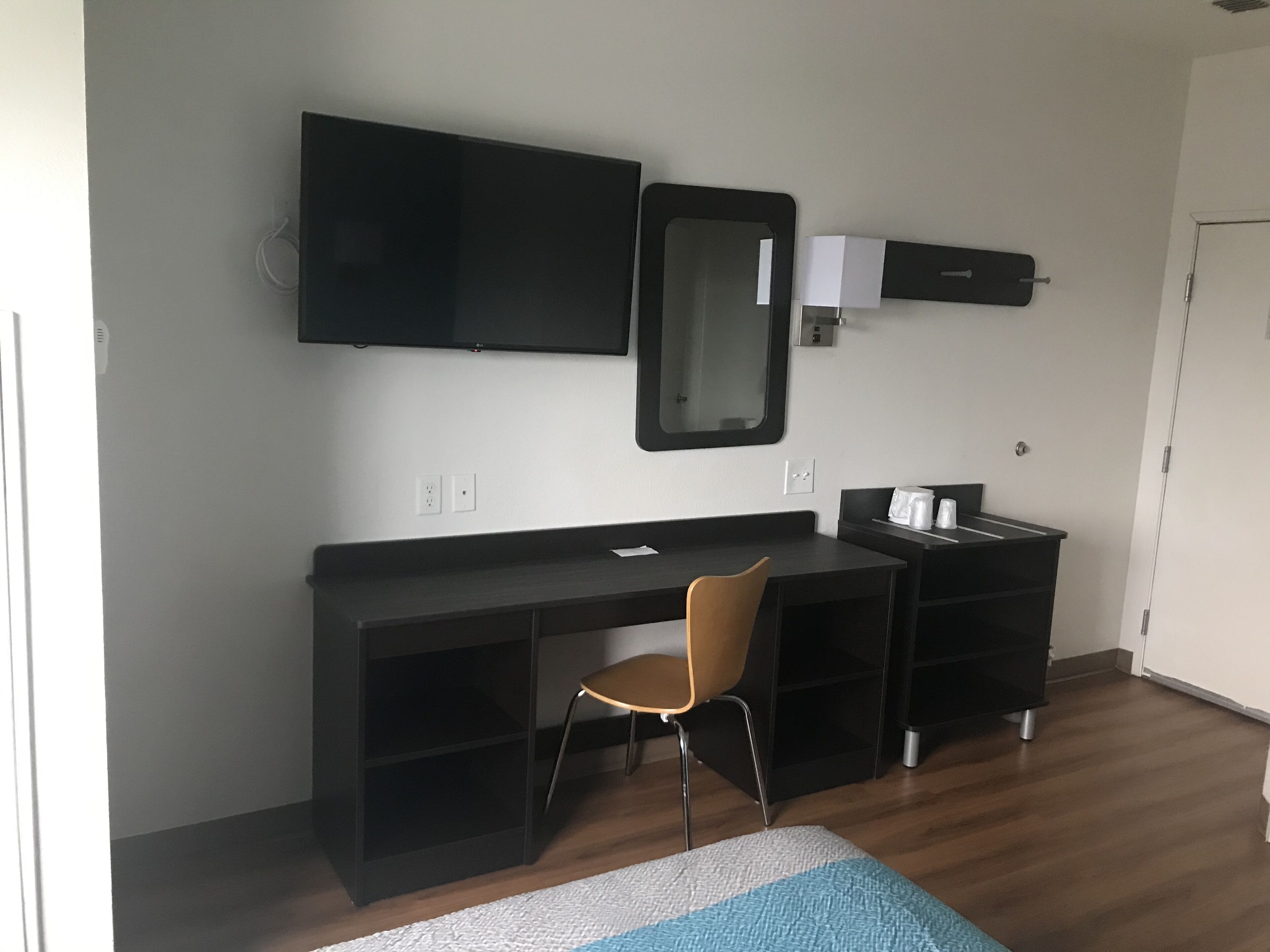A modern room with a dark wooden desk, a chair, a flat-screen TV mounted above the desk, a mirror, and a small cabinet with a coffee maker.