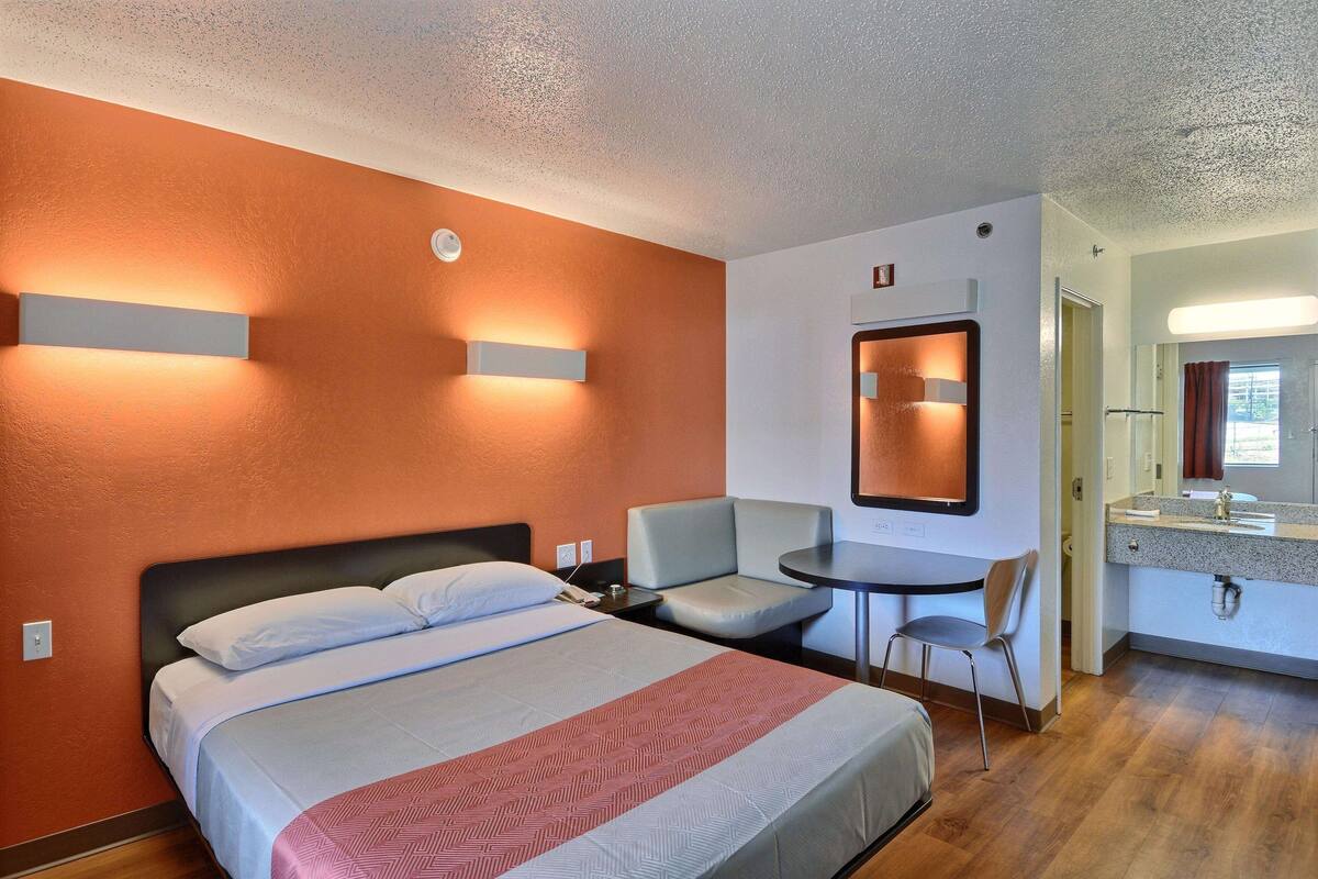 Hotellihuone, jossa on sänky, pieni pöytä, tuoli sekä kylpyhuone, jossa on pesuallas ja peili.
