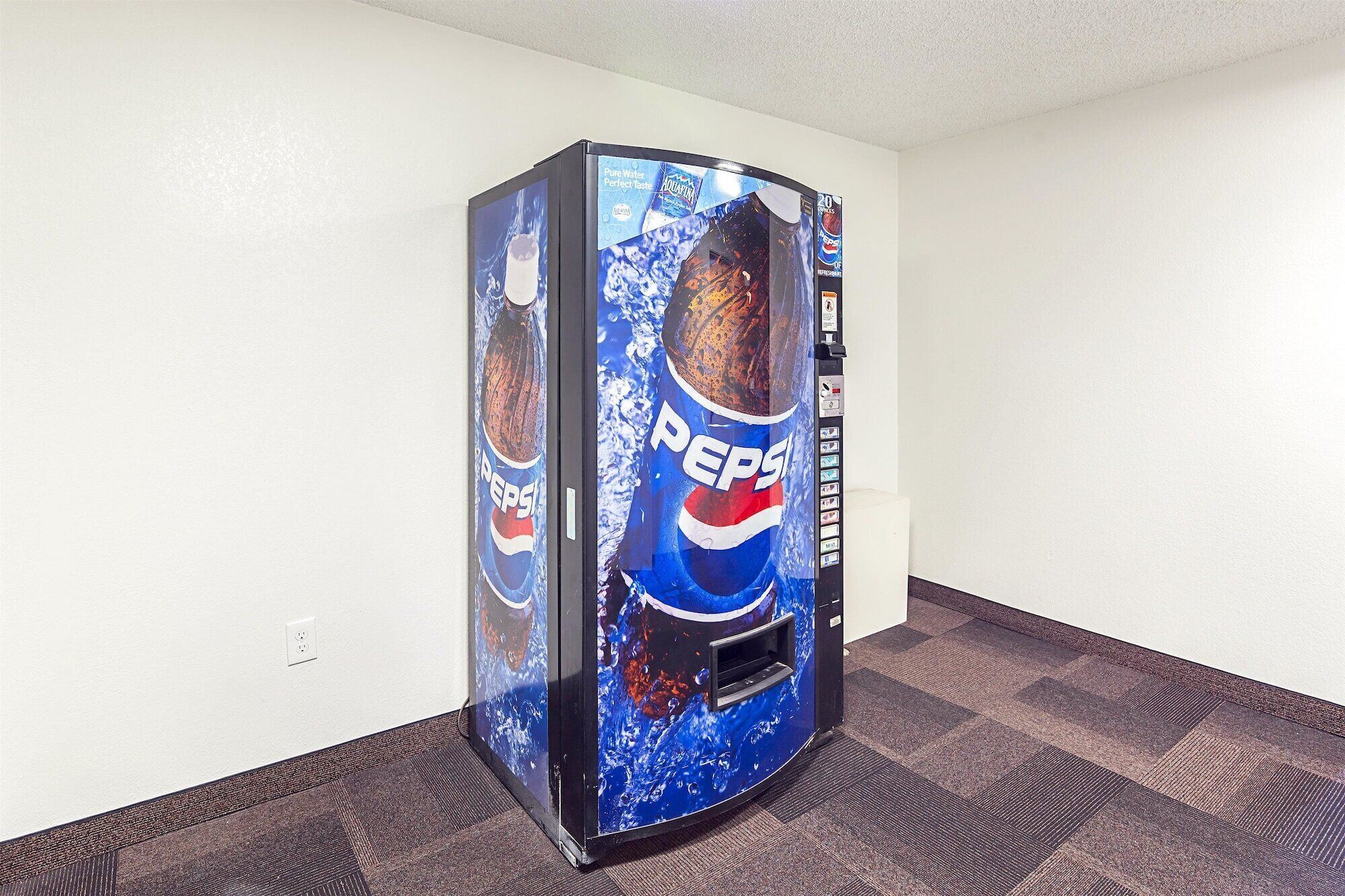A vending machine with a Pepsi logo, located in a room with a carpeted floor and white walls.