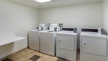 Une buanderie équipée de quatre machines à laver et de quatre sèche-linge à chargement frontal, tous de couleur blanche, d’une étagère blanche et d’un sol carrelé.
