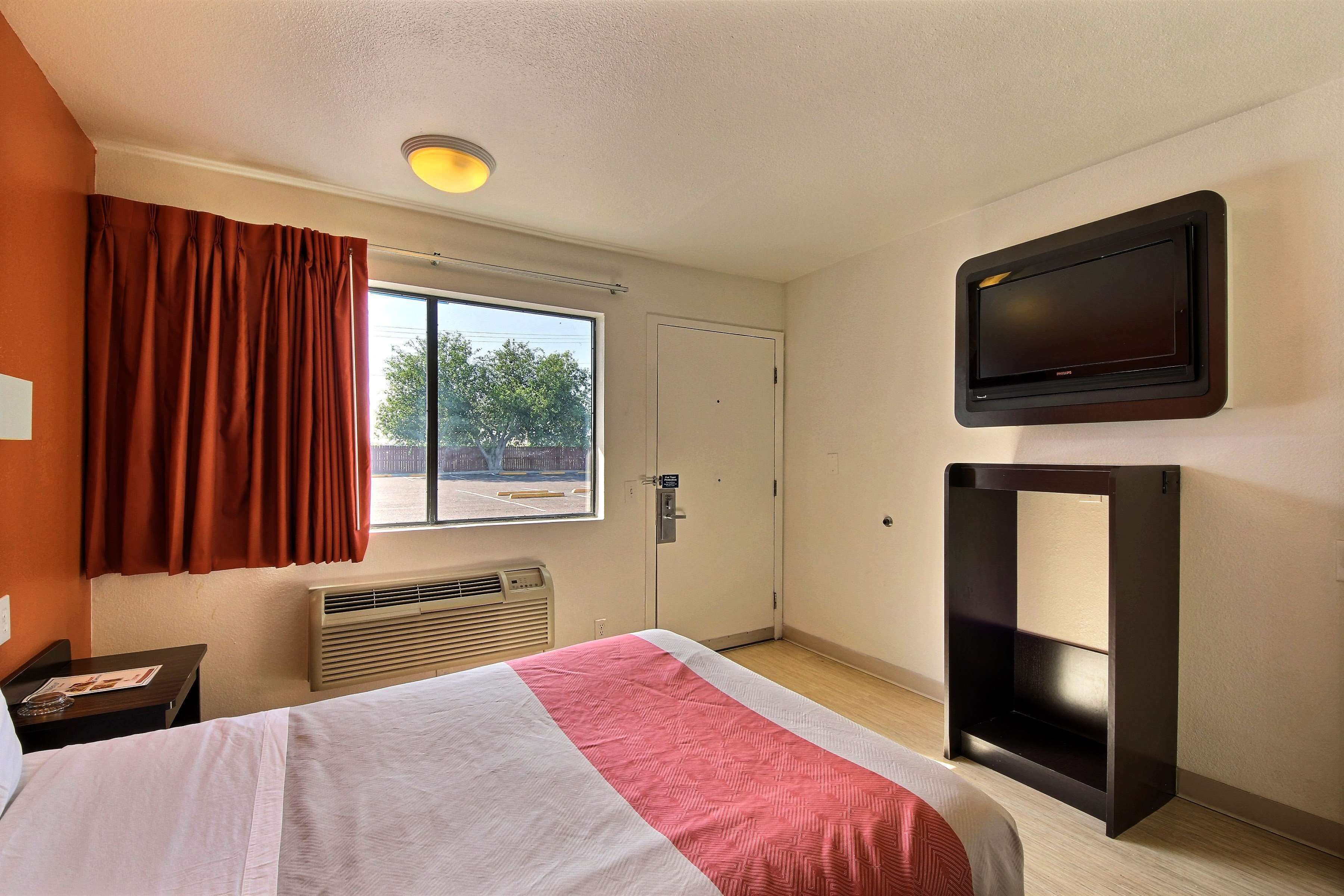Una camera d'albergo con un letto, una TV a parete, una finestra con tende, un termosifone e una porta.