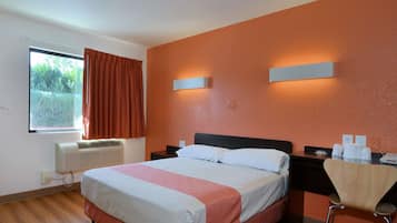 A hotel room with a bed, a desk, a chair, a window with curtains, and a wall-mounted light fixture.
