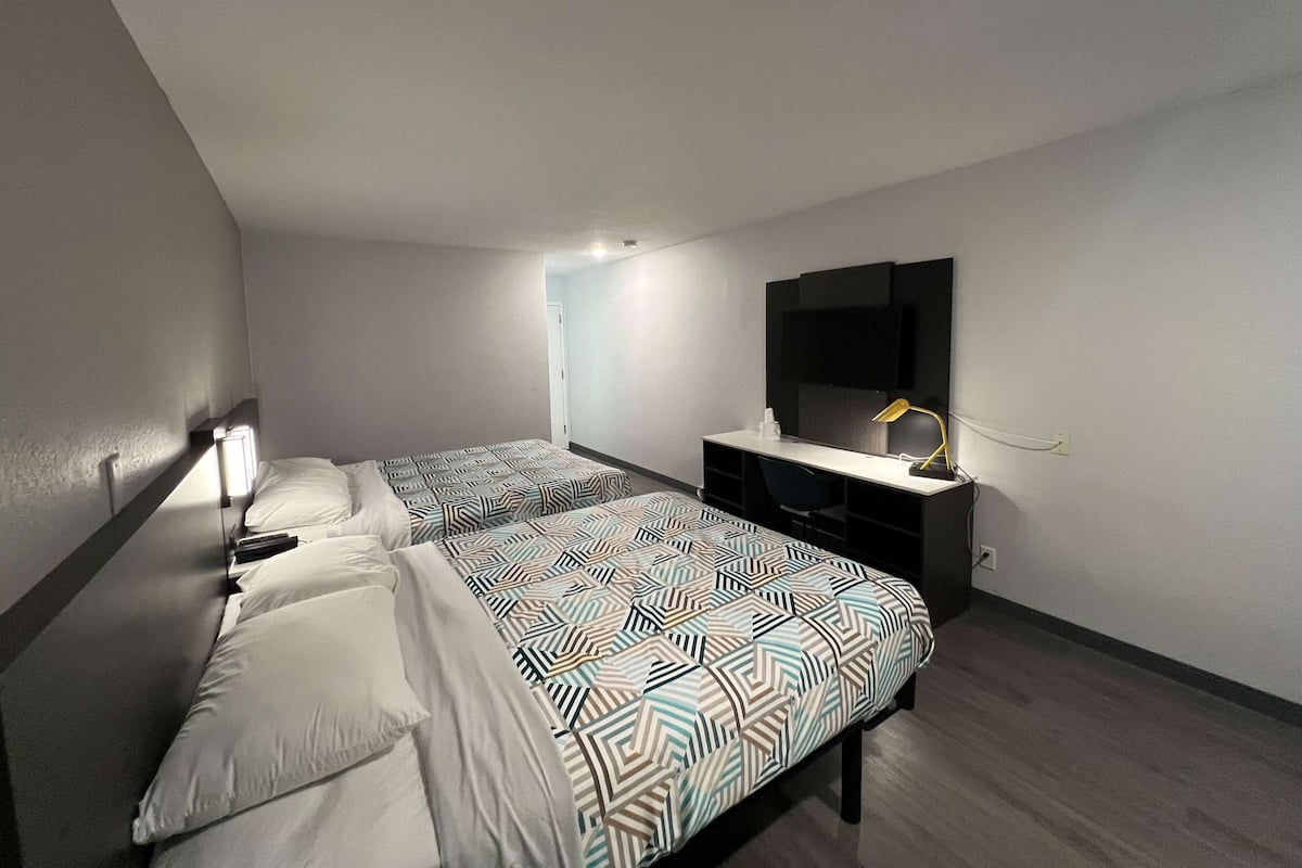 A hotel room with two beds, a desk, a chair, a TV, and a lamp.