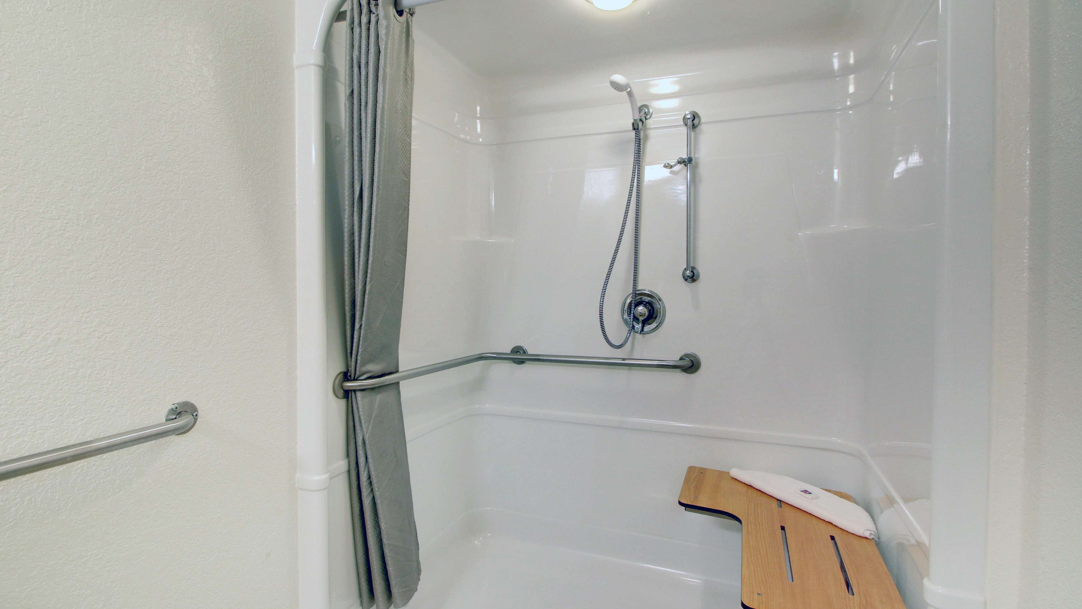 Zona de ducha con barra de apoyo, cabezal de ducha y un tapete de baño de madera.