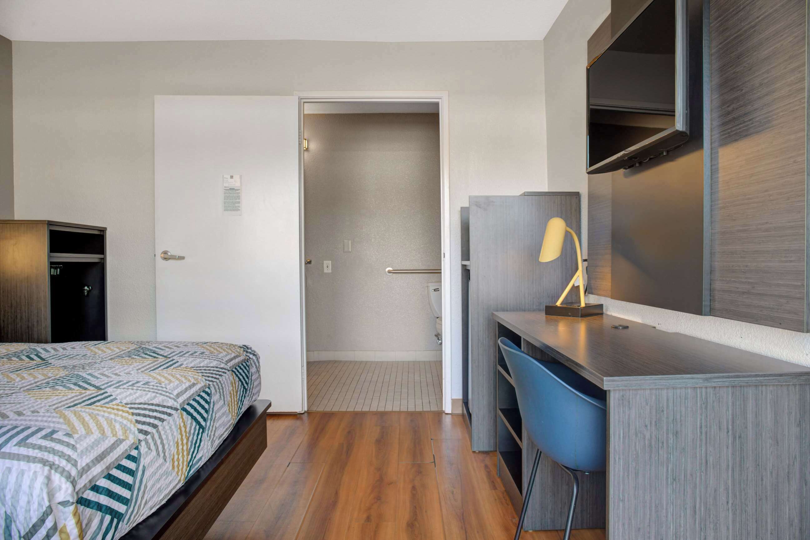 A hotel room with a bed, a desk, a chair, a TV, and a door leading to a bathroom.