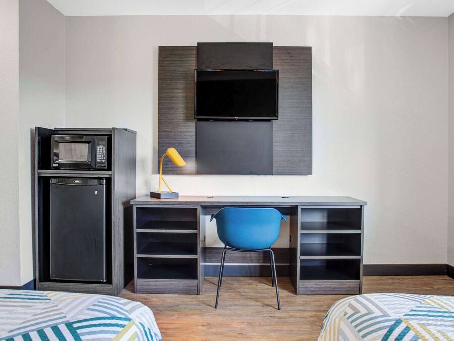 A compact hotel room with a bed, a desk with a chair, a microwave, and a TV mounted on the wall.