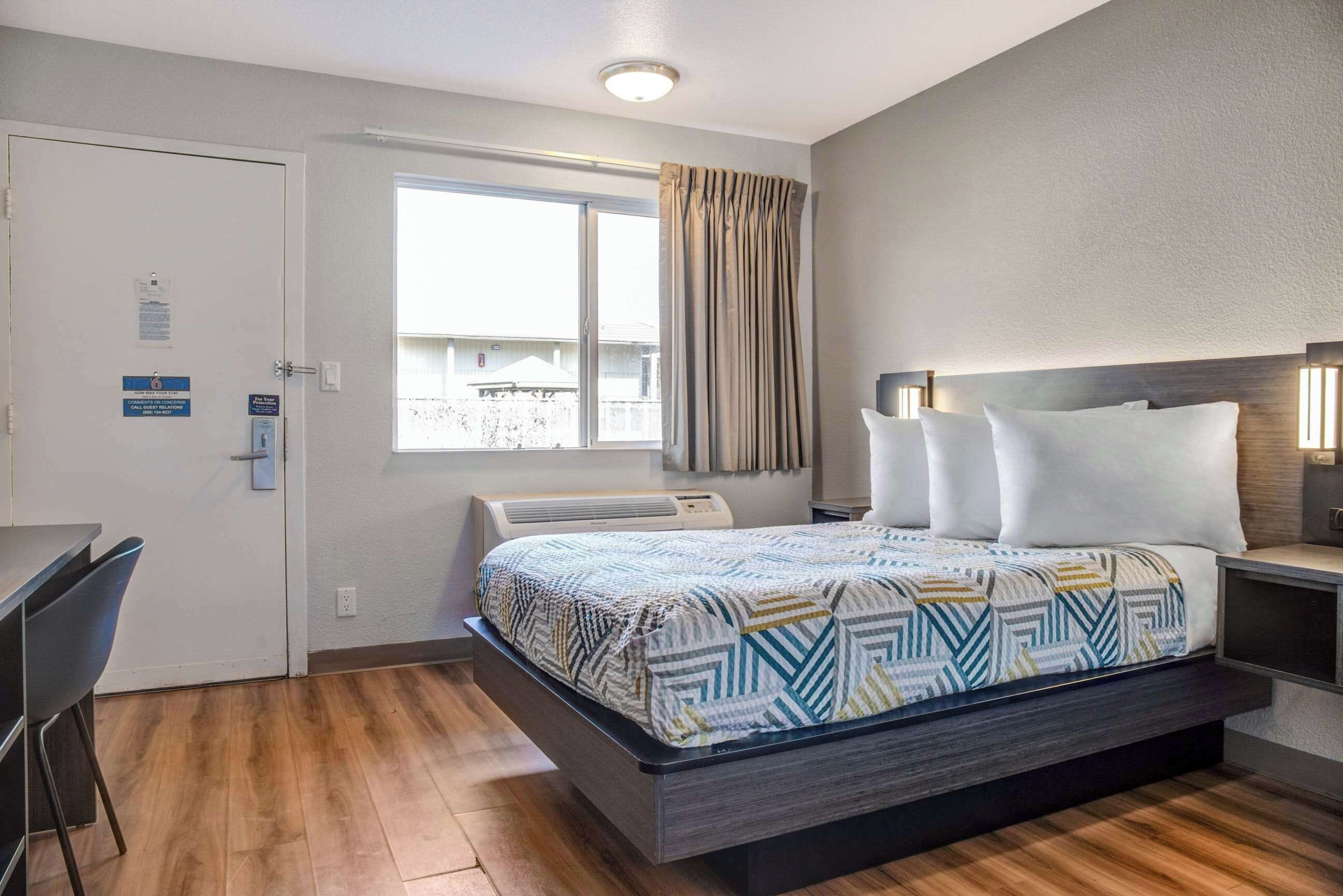 A hotel room with a bed, a desk, a chair, a window with curtains, and an air conditioning unit.