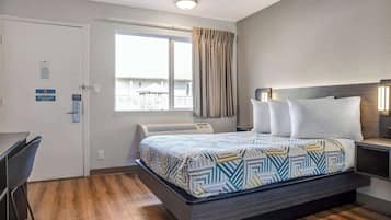 A hotel room with a bed, a desk, a chair, a window with curtains, and an air conditioning unit.