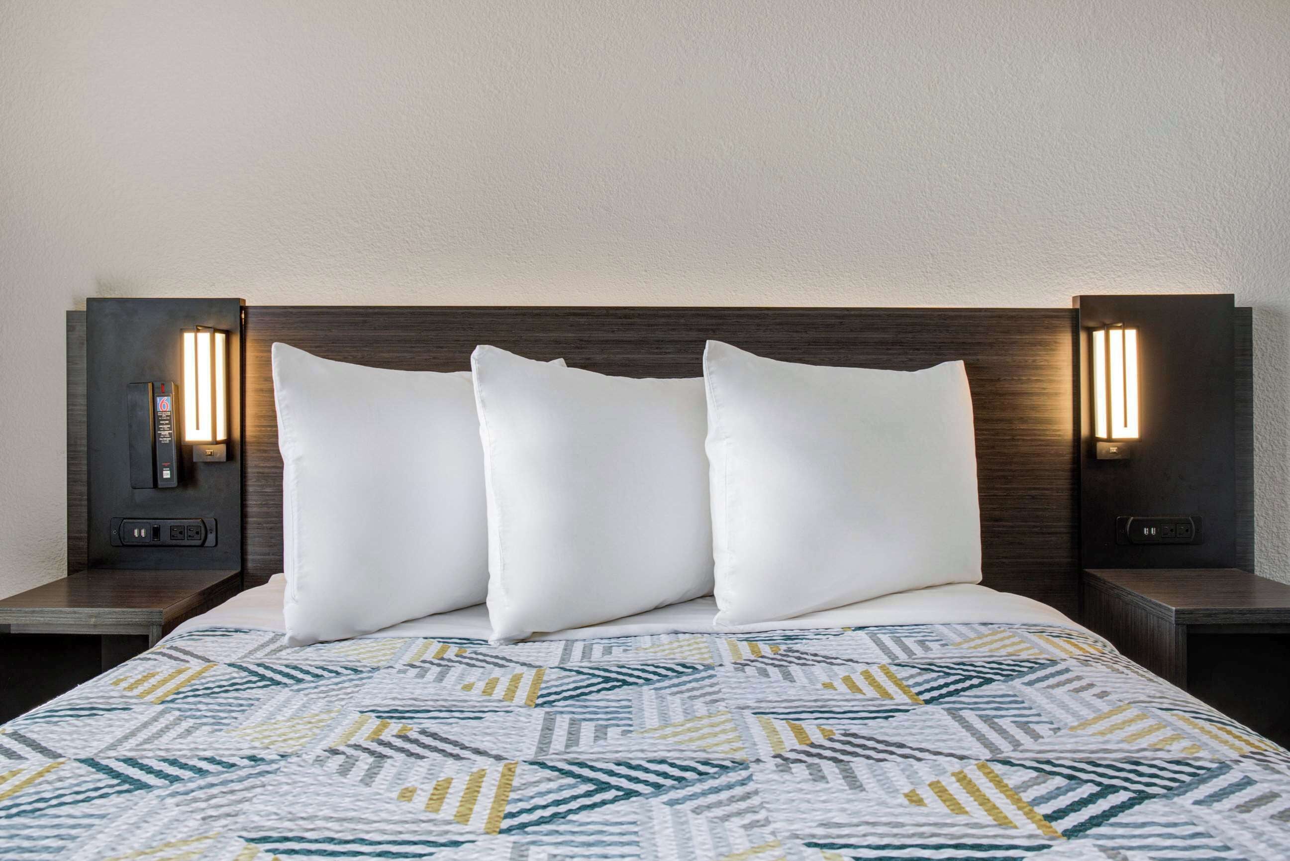 Quarto de hotel com cama, almofadas brancas, colcha estampada e iluminação fixada na parede.