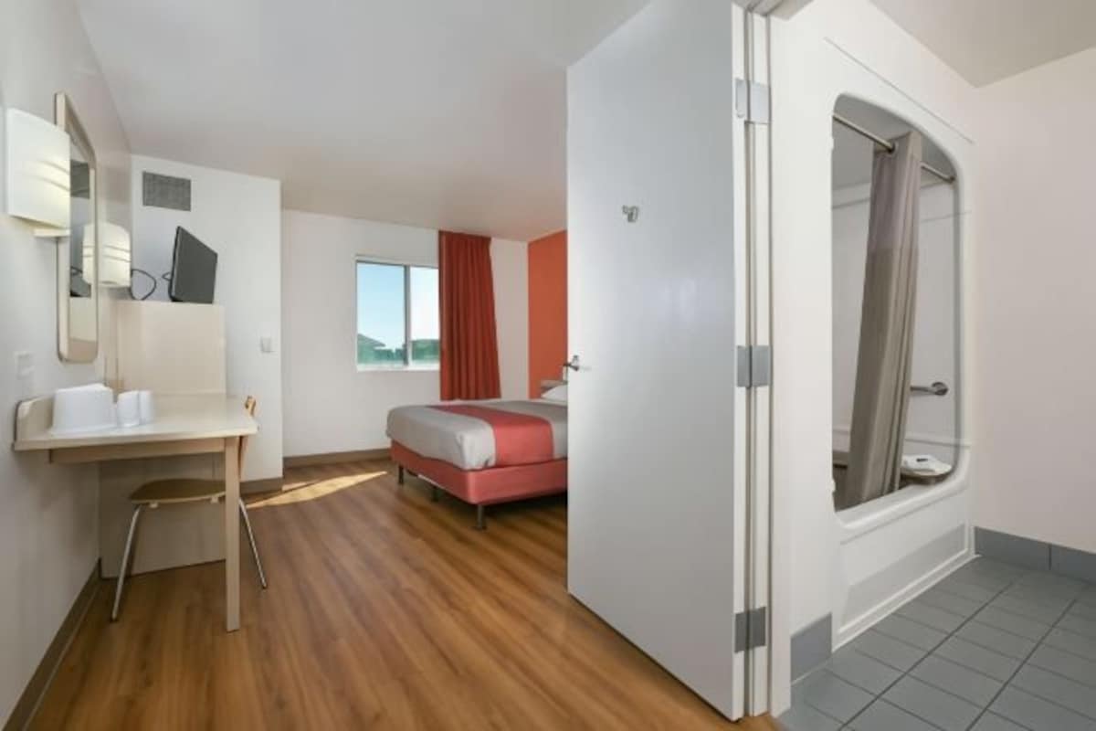 A modern hotel room with a wooden floor, a bed with red bedding, a desk with a chair, a large window with curtains, and a shower area.