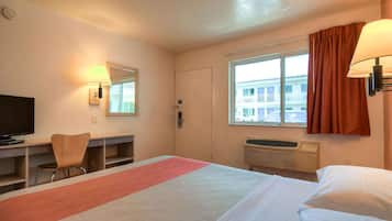 Habitación de hotel con cama, escritorio y silla, televisión, ventana con cortinas y aire acondicionado.