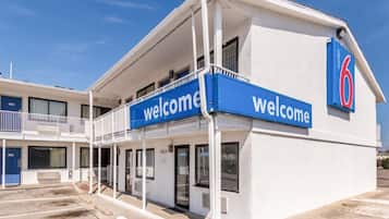 Um motel com um letreiro de 'Welcome', uma área de estacionamento coberta e céu claro.