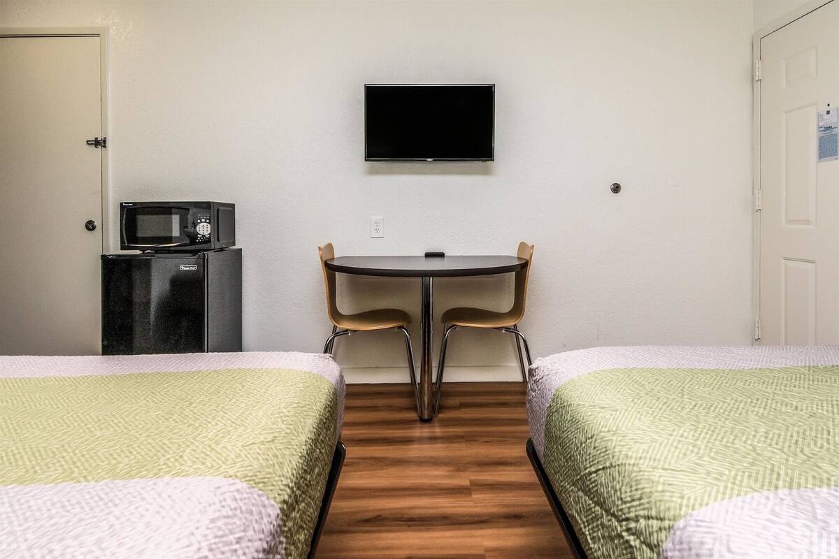 Ett rum med två sängar, ett litet bord med stolar, en mikrovågsugn och en tv som hänger på väggen.