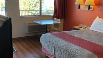 Et hotelværelse med seng, skrivebord, stol, et vindue med gardiner og en lampe monteret på væggen.