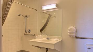 Un baño con ducha, lavamanos, espejo y dispensador de toallas.