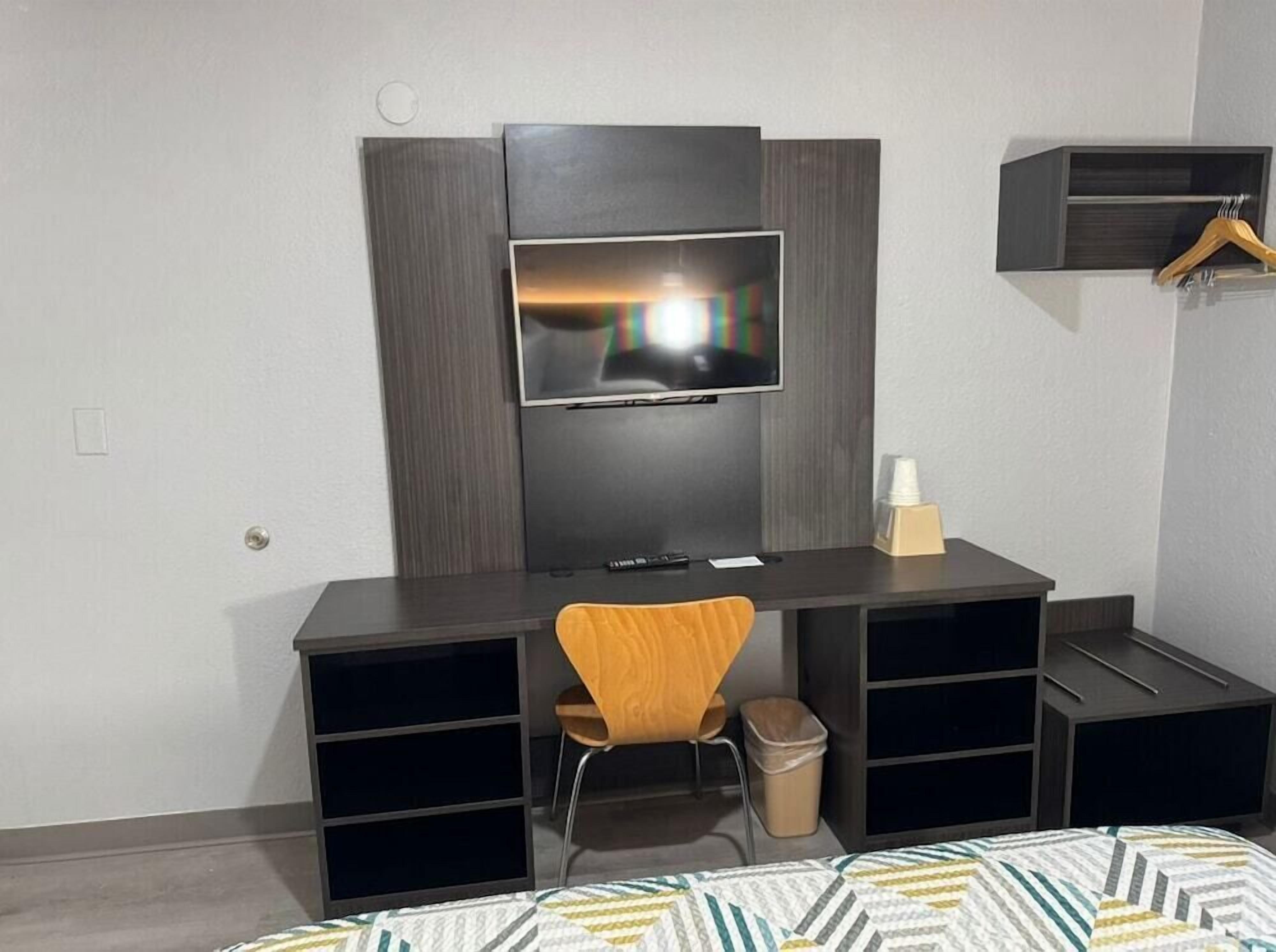 Una habitación de hotel moderna con un escritorio de madera oscura, un televisor de pantalla plana montado en la pared, una silla de madera y una cama con edredón estampado.