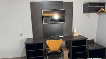 A modern hotel room with a dark wood desk, a flat-screen TV mounted on a wall, a wooden chair, and a bed with a patterned bedspread.