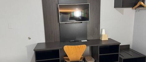 A modern hotel room with a dark wood desk, a flat-screen TV mounted on a wall, a wooden chair, and a bed with a patterned bedspread.