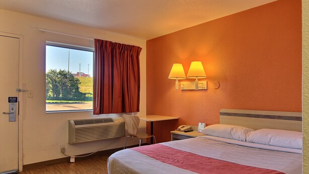 A hotel room with a bed, a desk, a chair, a window with curtains, a wall-mounted lamp, and a radiator. - Motel 6 Bismarck, ND (Bismarck)