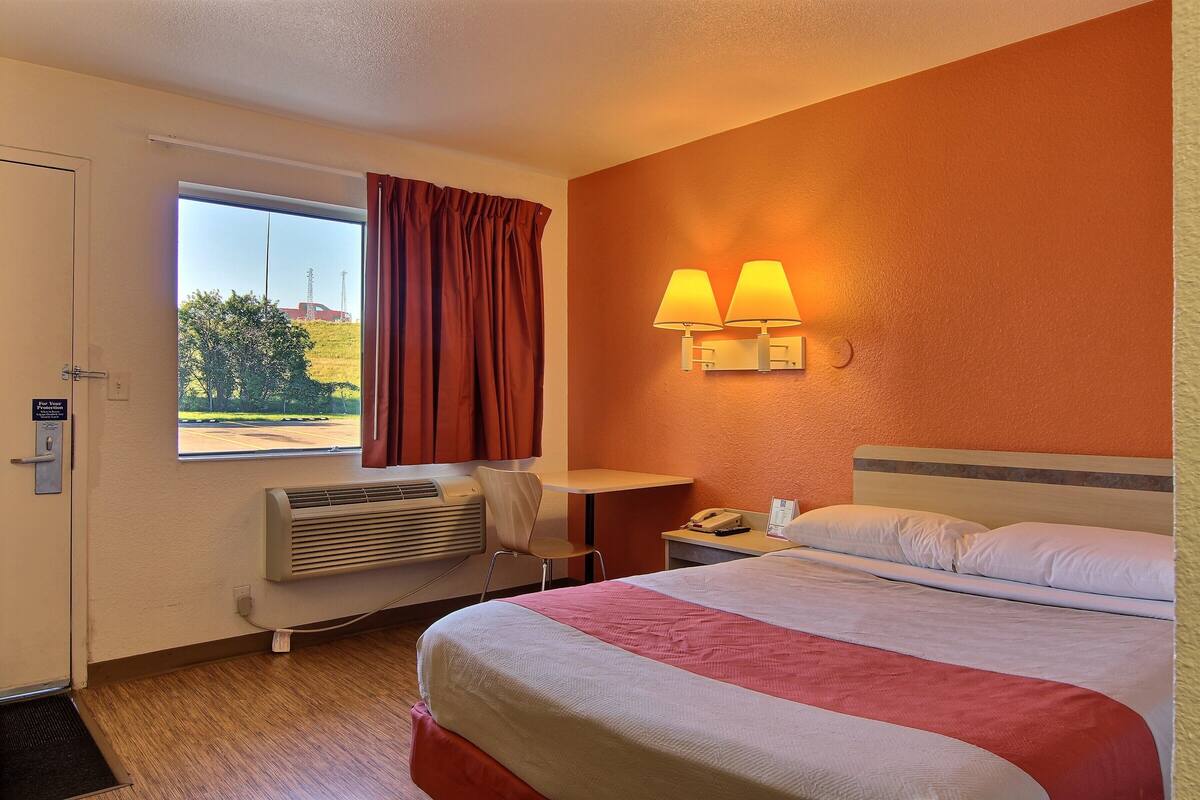 A hotel room with a bed, a desk, a chair, a window with curtains, a wall-mounted lamp, and a radiator.