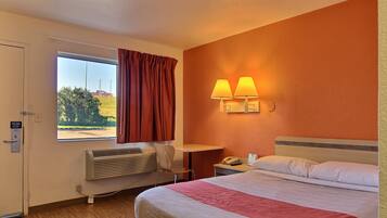 A hotel room with a bed, a desk, a chair, a window with curtains, a wall-mounted lamp, and a radiator.