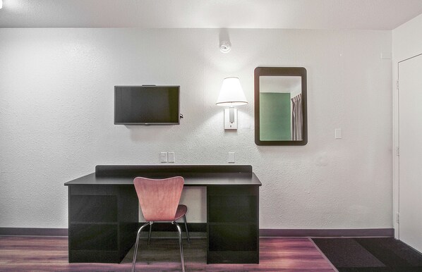 A small, modern bathroom with a dark vanity, a pink chair, a wall-mounted TV, a mirror, and a light fixture. - Motel 6 Green Bay, WI (Green Bay)