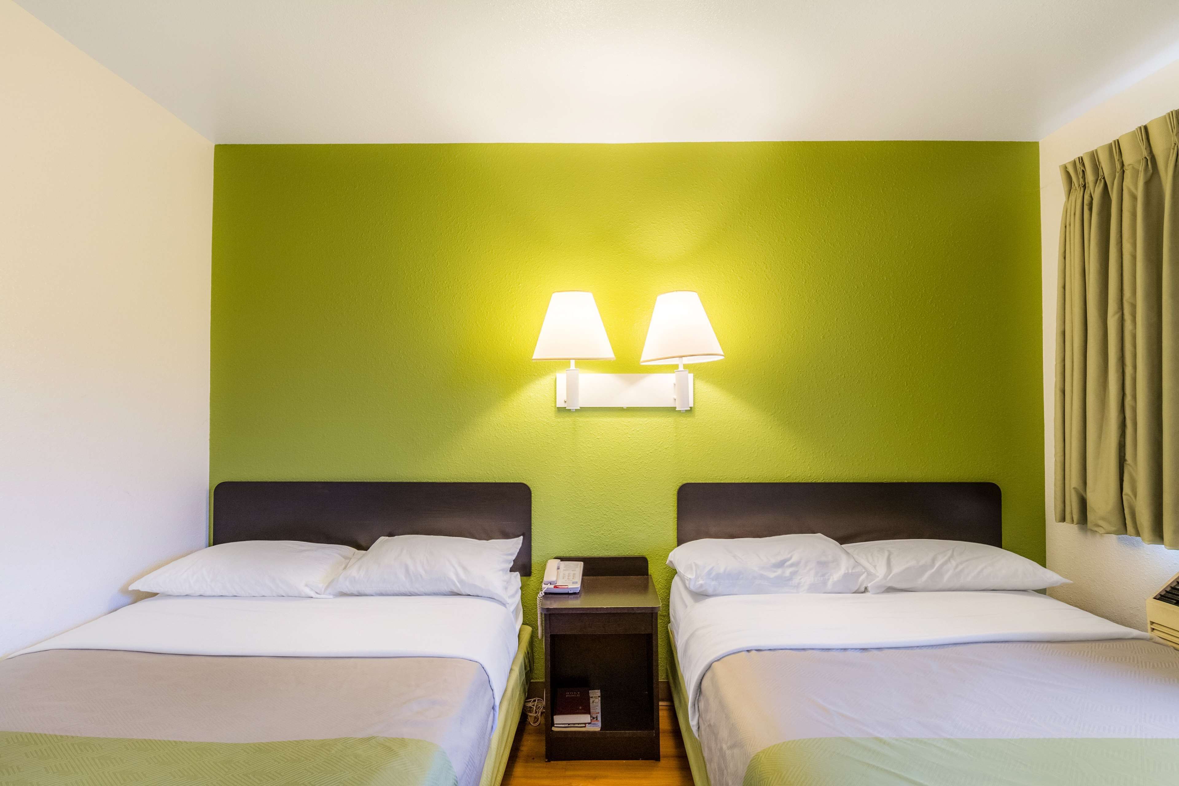 A hotel room with two beds, a nightstand with a lamp, and a green wall.