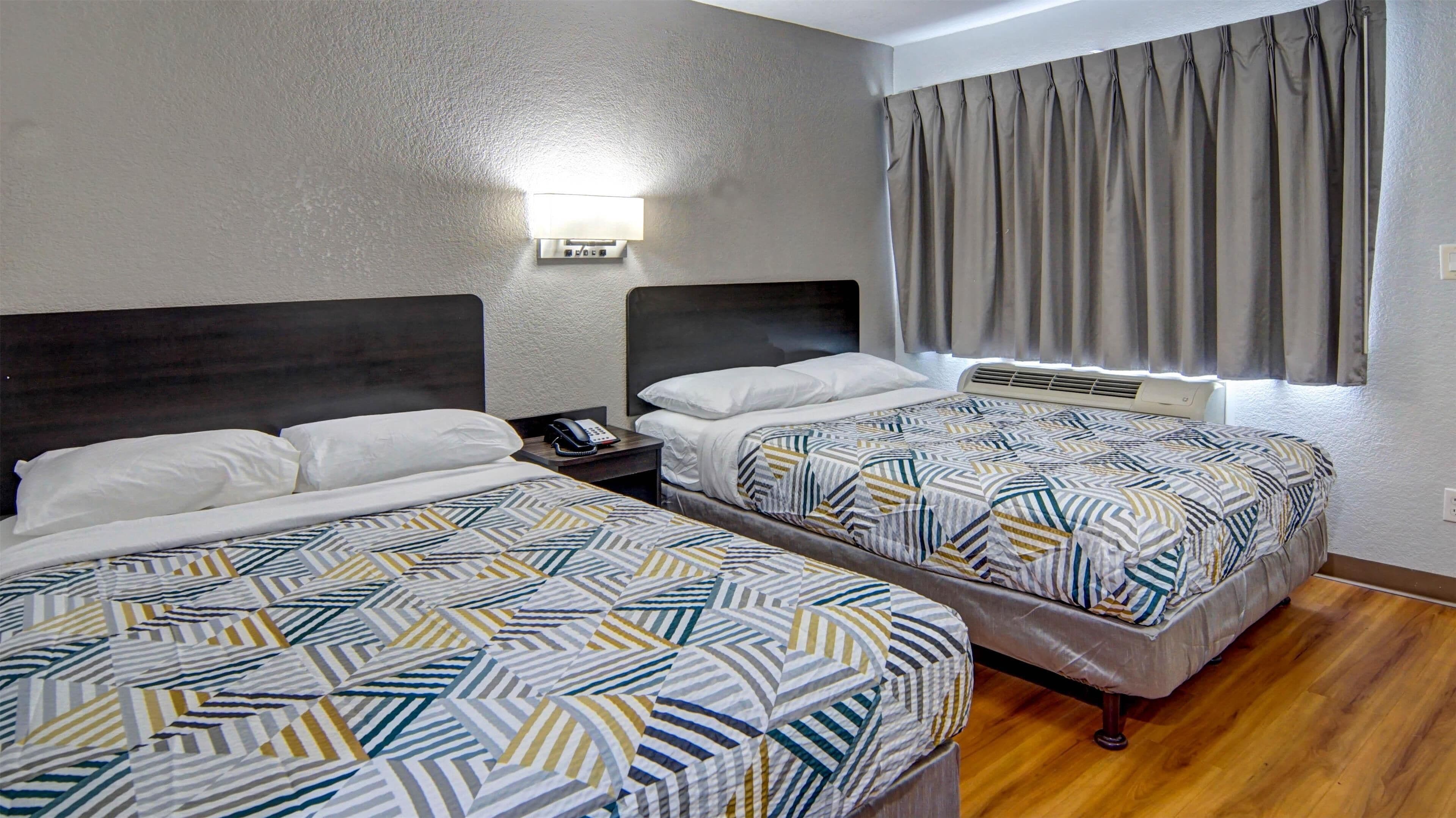 A hotel room with two beds, a nightstand with a phone, a wall-mounted lamp, and a window with curtains.