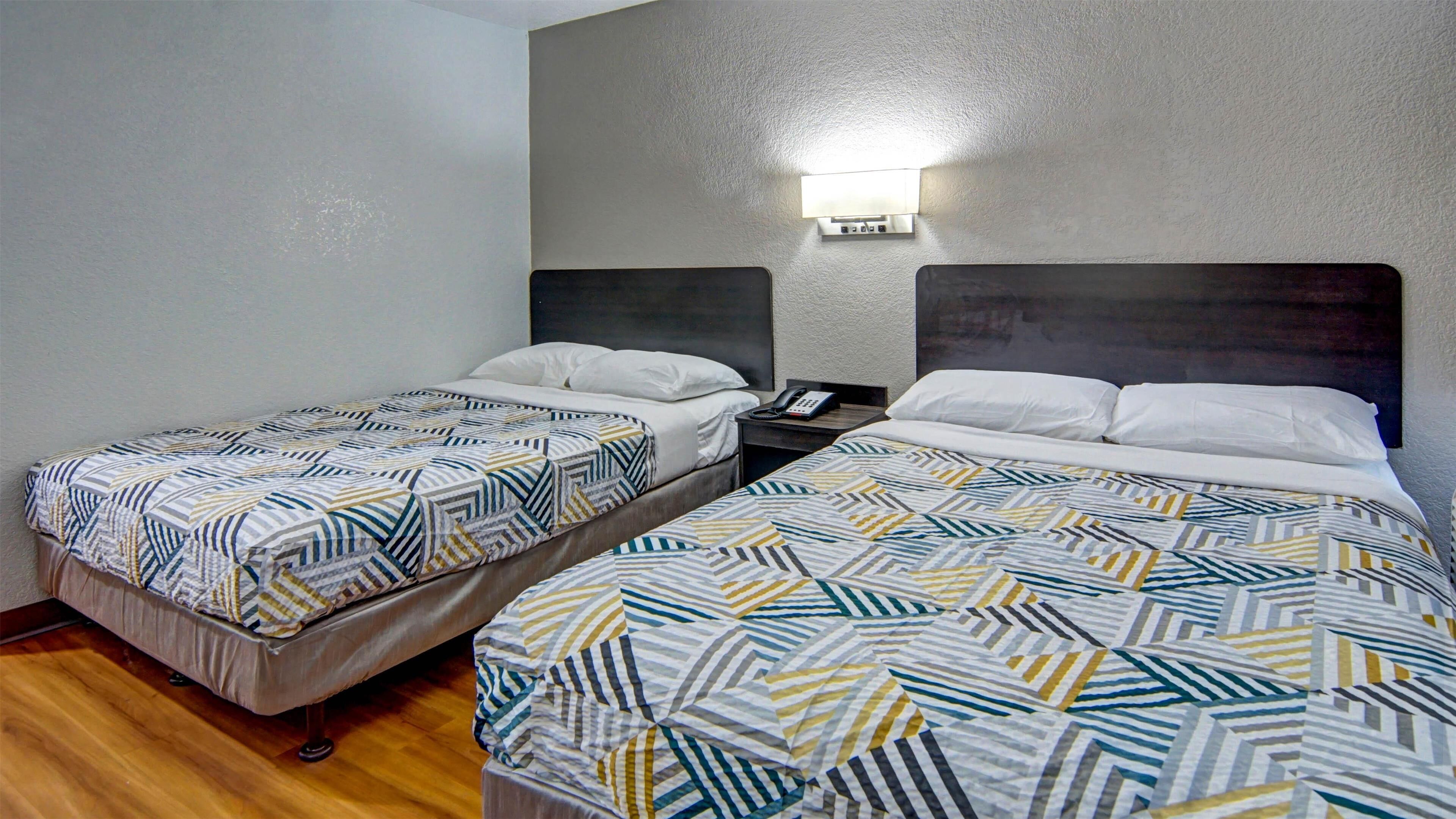 Habitación de hotel con dos camas individuales, cada una con colcha estampada y almohadas blancas. Se observa una mesita de noche con un teléfono y una lámpara en la pared.