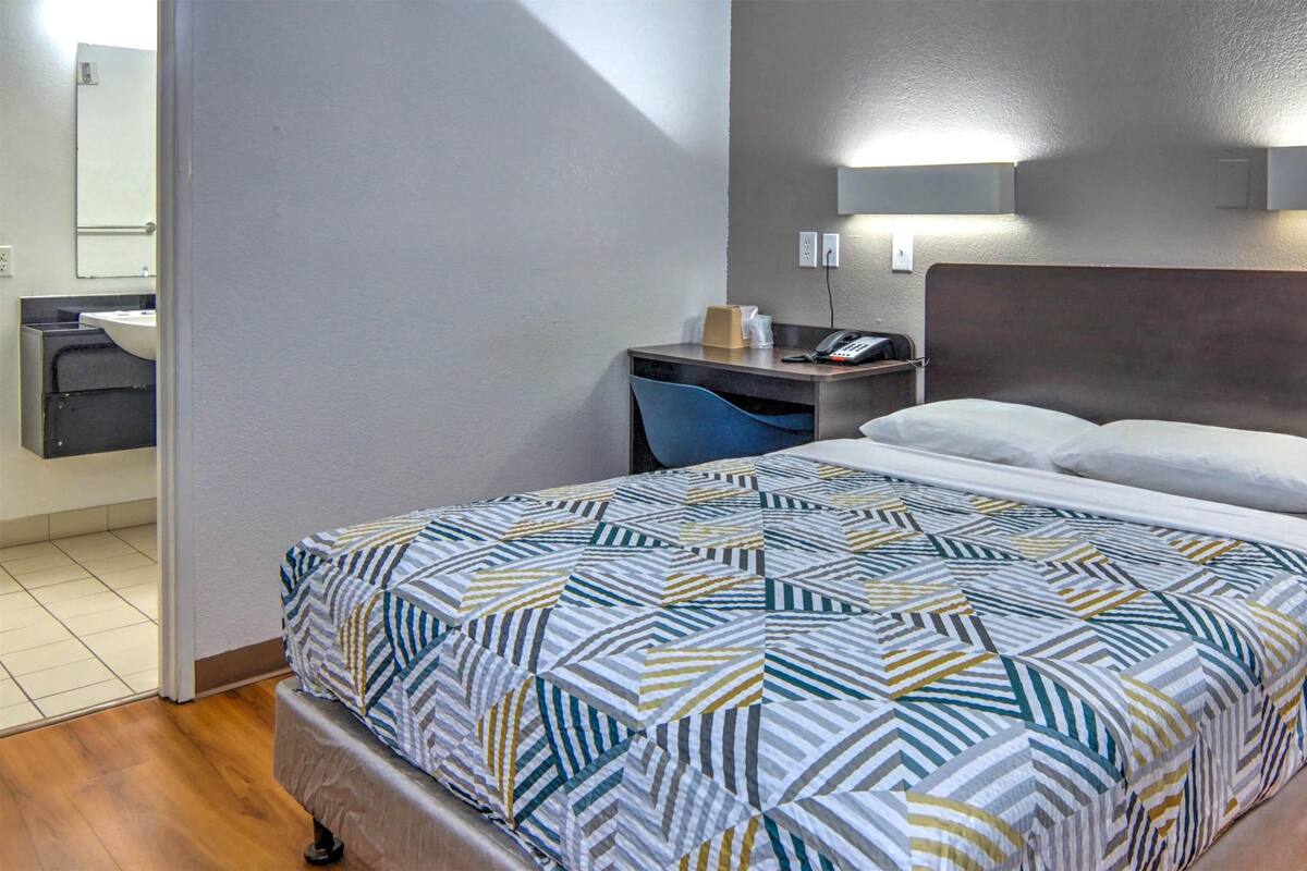 A hotel room with a bed, a nightstand, a desk, a chair, and a bathroom visible through an open door.
