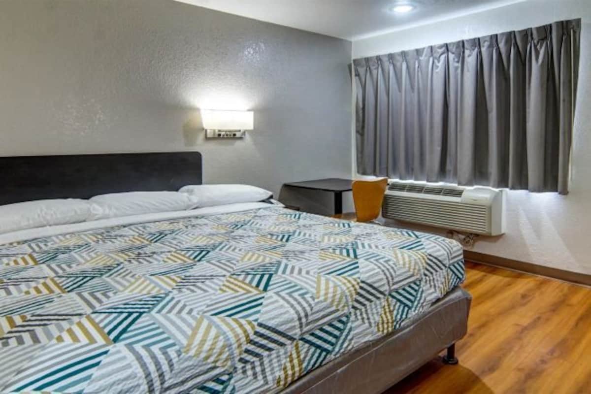 A hotel room with a large bed, a bedside table, a lamp, a window with curtains, and an air conditioning unit.