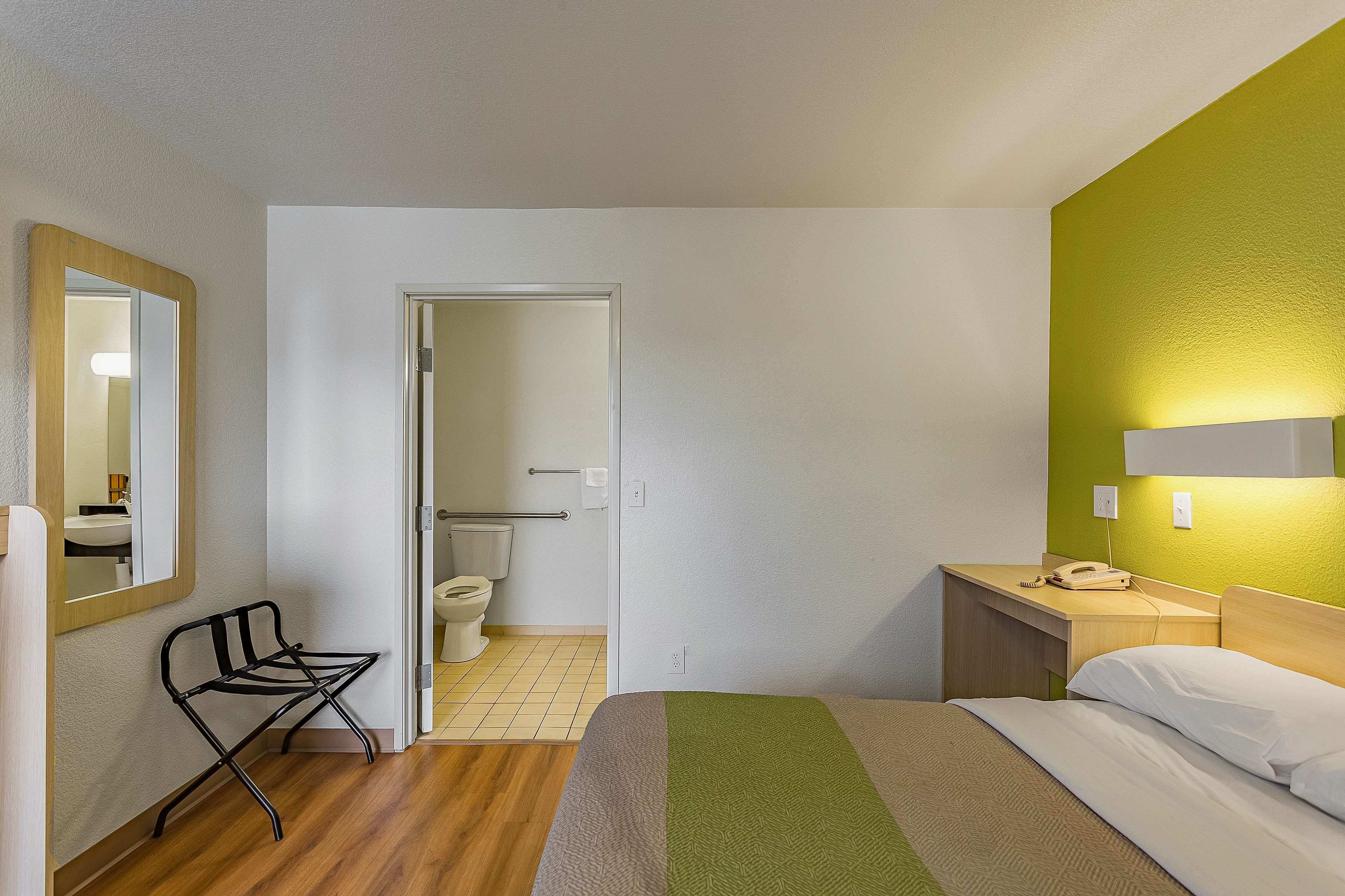 A hotel room with a bed, a bedside table with a lamp, a chair, and a bathroom visible through an open door.