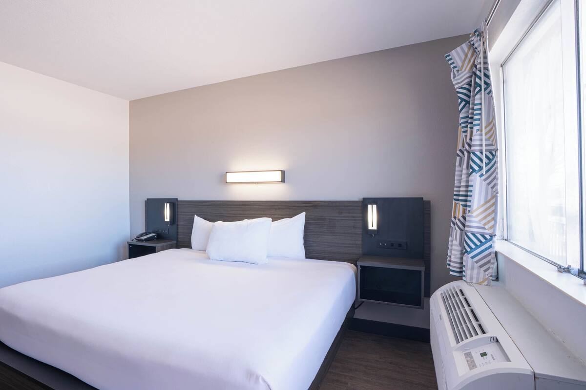 A hotel room with a large bed, bedside tables, a wall-mounted light, and a window with curtains.