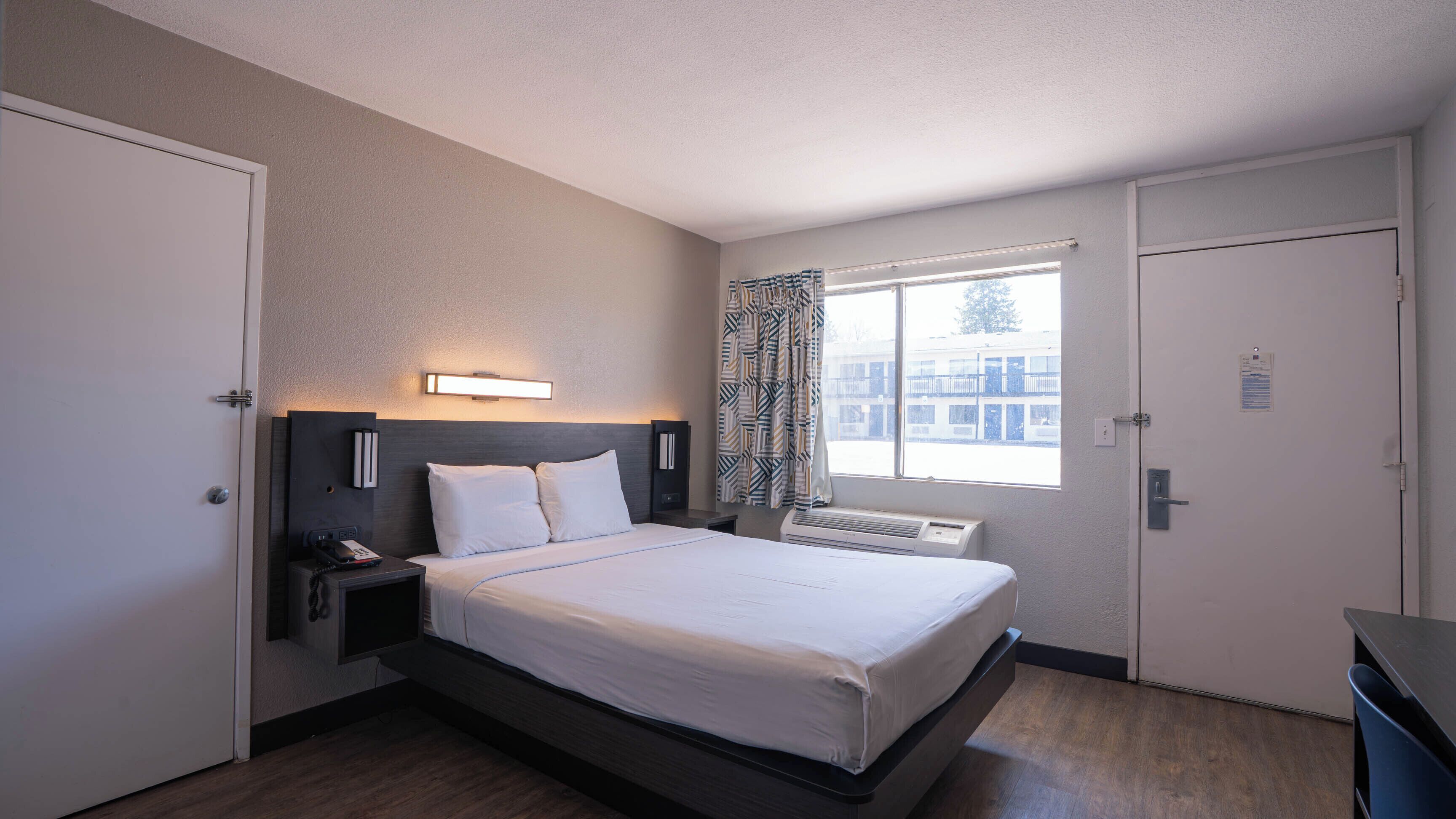 A hotel room with a bed, bedside table with a phone, a wall-mounted light, a window with curtains, and a radiator.