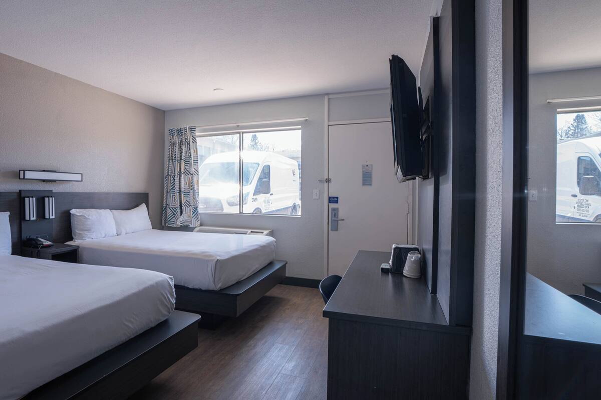 A hotel room with two beds, a desk, a TV, and a window with a view of a white van outside.